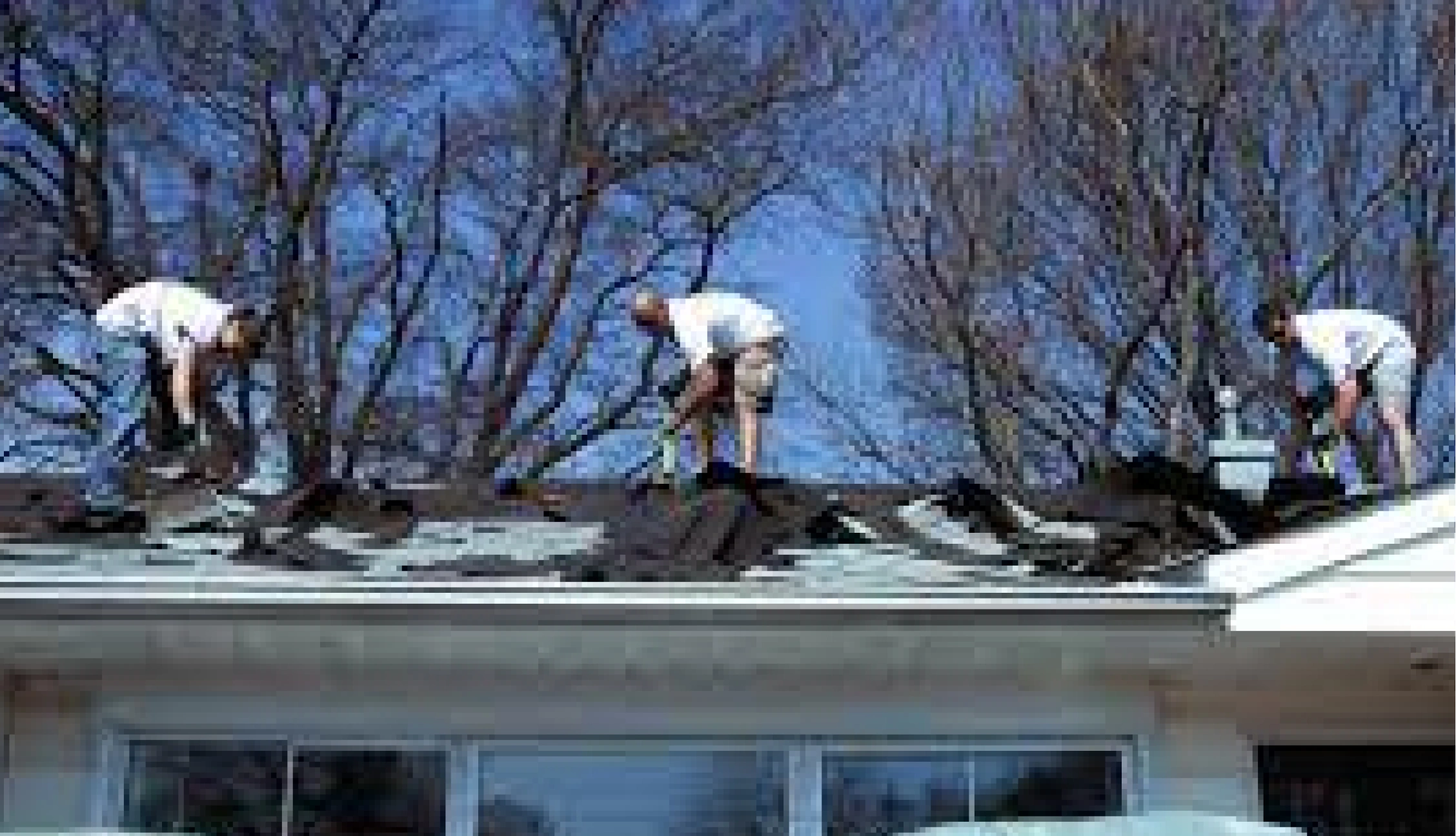 Three men installing new black shingles on a white house with many trees in the background